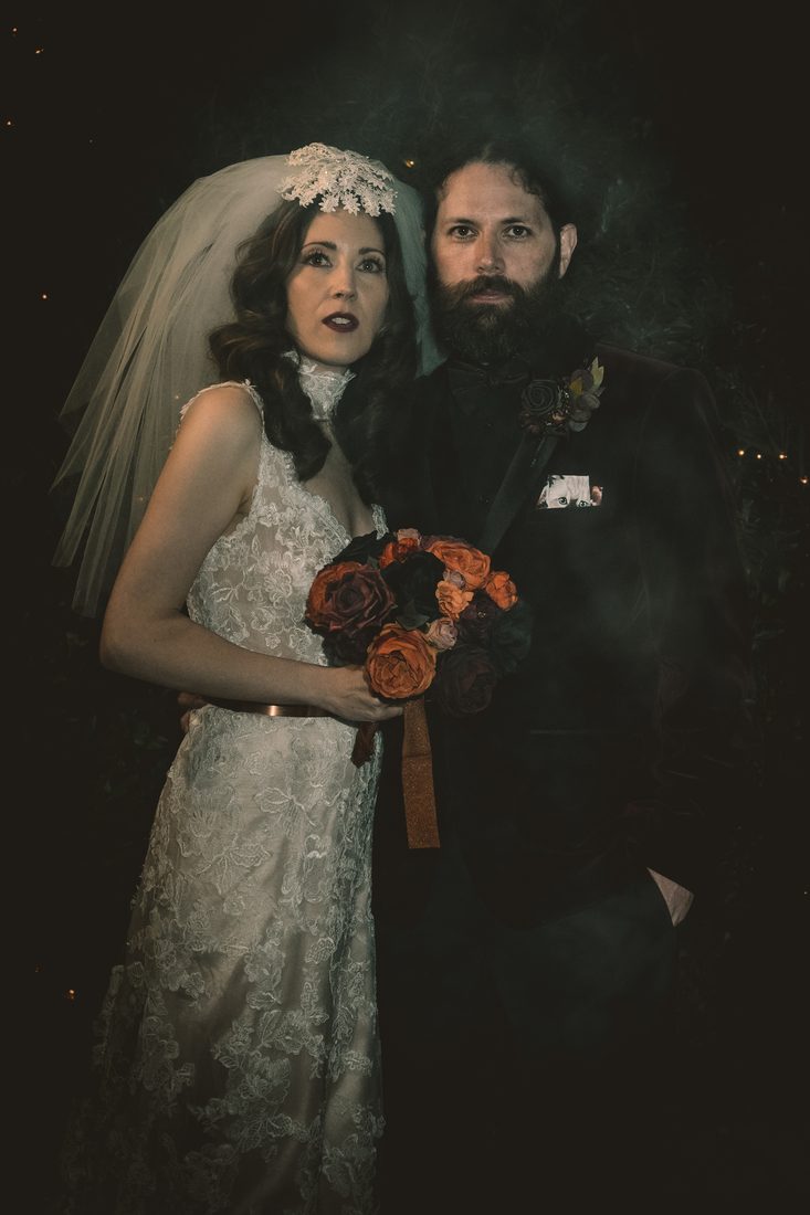 Sean and Bethany on their wedding day — she in a lace gown and veil holding a bouquet of orange and black roses, he in a dark suit with a black rose boutonnière